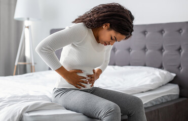 Portrair of upset black woman suffering from strong abdominal pain, touching her tummy, sitting on...