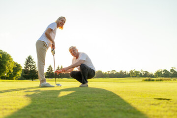 elderly senior couple in uniform playing golf on golf course at sunset, old man instructor teaching...