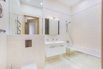 A white tiled bathroom with mirrored surfaces, a large bathtub and a sink.