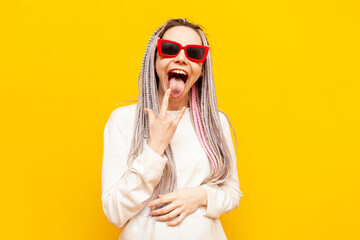 young girl with colored dreadlocks and sunglasses shows rock gesture and screams on yellow isolated background, crazy hipster woman with unique hairstyle and braids shows tongue