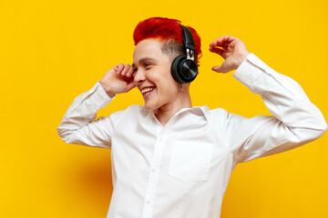 young short-haired red-haired girl listens to music on headphones and dances on a yellow isolated background, hipster woman with piercing moves with her arms raised and sings