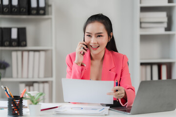 Confident Businesswoman on the Phone: A smiling young woman in a bright pink blazer confidently makes a phone call while reviewing documents at her desk in a modern office.