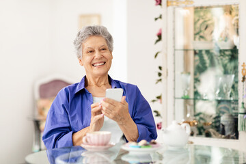 A happy elderly woman sits at a table in a bright cafe, using her smartphone. She is enjoying coffee and desserts, embracing a relaxed retirement lifestyle. Friends are likely nearby.