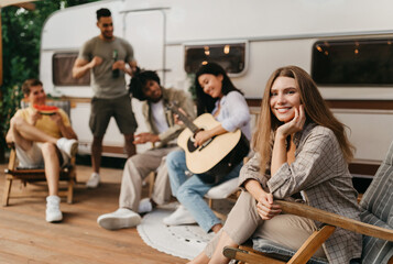 Group of happy young people having fun on camping trip, sitting in lounge chairs, playing guitar, drinking beer, eating watermelon, enjoying cool summer vacation in countryside, copy space