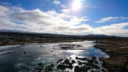 Iceland - 3.24.2018: A partly frozen river among dried meadows and winter trees with snow mountains at the horizon under a cloudy blue sky with sunlight in Thingvellir National Park before pandemic