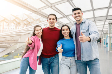 A cheerful group of two couples poses together at the airport, smiling and giving thumbs up as they prepare for their weekend getaway. The indoor space is bright and welcoming.