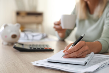 Fototapeta premium Budget planning. Woman with cup of drink taking notes at table indoors, closeup
