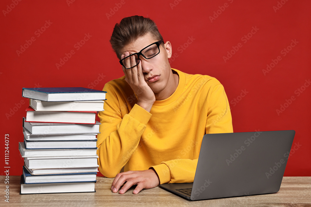 Poster tired student before exam with stack of books and laptop at wooden table against red background