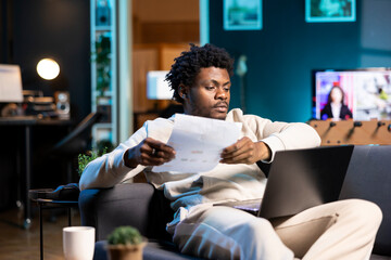 Man at home paying attention in elearning seminar economy class, looking at auxiliary school materials. Homeschooling student lounged on couch in living room listening to lesson and checking files