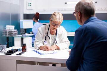 Senior doctor sitting at desk and writing sickness symptoms of male pensioner patient during medical checkup in hospital. Elderly female physician using clipboard at clinic consultation with old man.