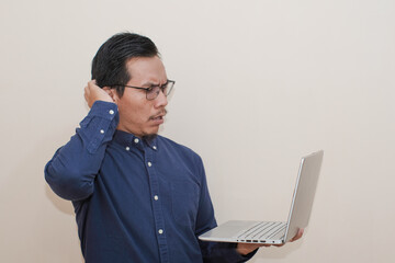 Shocked Southeast Asian man wearing blue shirt with glasses holding laptop, standing over isolated white background, Positive person