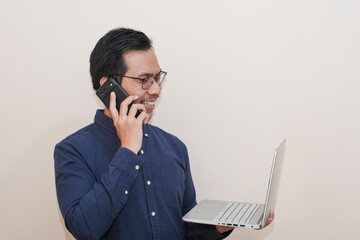 Smiling Southeast Asian man wearing blue shirt with glasses holding laptop and using smartphone taking a call, standing over isolated white background, Positive person