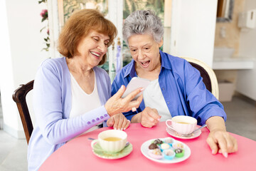 Two elderly women share a lighthearted moment at a cafe, looking at a smartphone together. They have tea and dessert pastries on the table, enjoying each other's company in retirement.