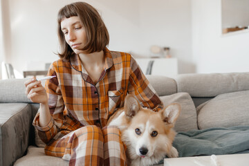 Sick Young Woman In Pajamas Sitting On A Sofa With Her Dog And Checking Her Body Temperature
