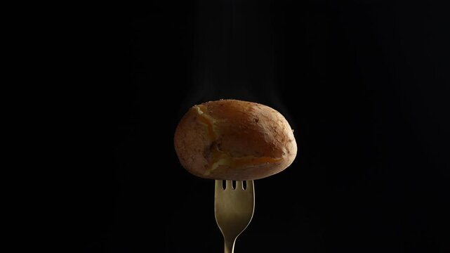 Fork with hot boiled potato on black background, close up, rotates. Jacket potato with cracked skin