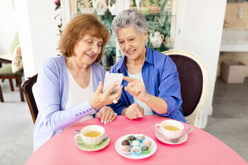 Two elderly women sit together at a round table enjoying dessert and tea. They are sharing laughter while looking at a mobile device. The cafe setting has a cozy ambiance.