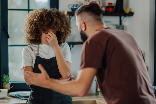 Young couple arguing in kitchen, woman touching face frustrated by man