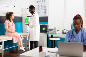 Physician holds tablet with greenscreen explains prenatal care with pregnant white woman while...