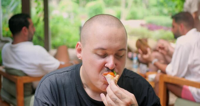A Caucasian man taking a bite of a delicious vegan pizza in a charming cafe in Bali. 