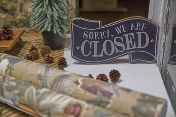 A shop window display with a 'Sorry, We Are Closed' sign, surrounded by pinecones and decorative paper rolls. A small artificial pine tree is also visible, creating a cozy atmosphere.