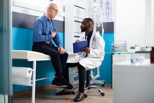 Caucasian old man mentioning his symptoms to african american physician during medical consultation. Young doctor with clipboard listening to health concerns from male pensioner patient.