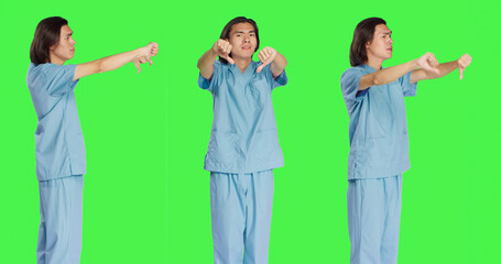 Obraz premium Asian specialist showing thumbs down symbol in studio, giving dislike and negativity gesture over greenscreen backdrop. Displeased medical nurse expressing disagreement, disapproval.