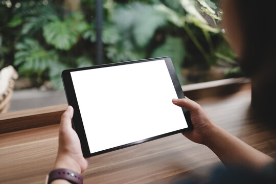 Hands holding black tablet device with blank white screen against wooden desk background, surrounded by green tropical plants, creating modern minimalist workspace atmosphere. - Powered by Adobe