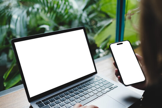 Person using laptop and smartphone with blank white screens against tropical plant background, showcasing modern devices for digital workspace mockup in natural environment setting.