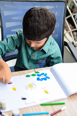 Smart Indian little boy perform thumb painting with different colourful water colour kit during the summer vacations, Cute Indian Kid doing colourful thumb painting drawing on wooden table