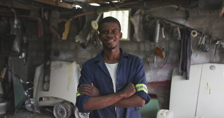 Portrait of a happy African male panel beater in a township workshop, looking at camera with arms cr