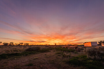 A glowing orange sunset viewed across a scrub and tree covered plain with farm trailers and equipment to one side with pink clouds in the darkening blue sky at Wilcannia in New South Wales, Australia.