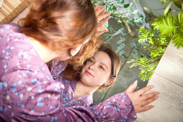 A woman in a floral purple dress gazes into a mirror surrounded by greenery in a tranquil indoor setting