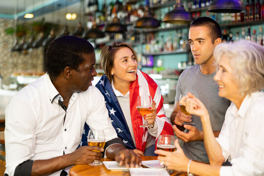 Young and senior football and soccer fans drinking beer at pub, cheering and celebrating scores. Girl with american flag on her shoulders - Powered by Adobe