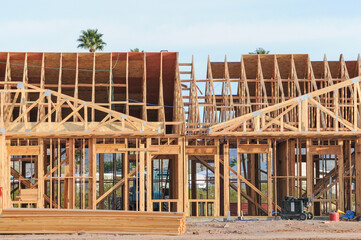 Wood-frame multifamily housing under construction with exposed trusses, OSB sheathing, and stacked lumber on site, illustrating early framing stage of residential development