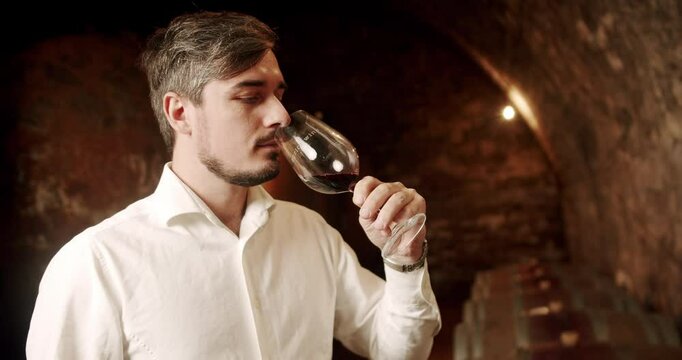 Man tasting wine in a factory or warehouse. Sommelier checking and drinking a glass of red wine. Sommelier admiring the wine in the old cellar. Male winemaker is tasting a red wine in a cellar.