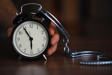 Hand Holding Alarm Clock with Handcuffs on Wooden Table Symbolizing Time Constraint and Restriction