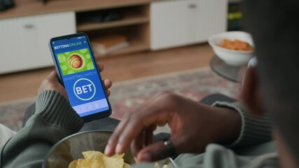 Over shoulder shot of Black man taking snacks from bowl while betting for basketball match using online gambling app on his smartphone, sitting on couch in front of TV