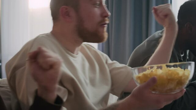 Three diverse male friends enjoying exciting sports game on TV, sitting on couch with bowl of snacks and props, including cheering sticks and wooden spinning ratchet noise maker grogger
