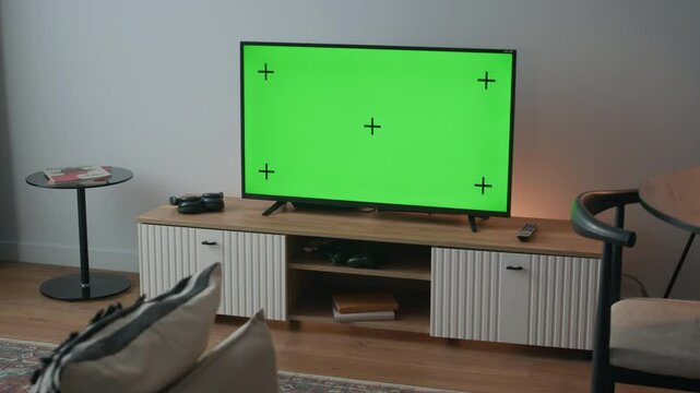 No people shot of modern living room with television displaying green screen with tracking markers on wooden console, flanked by game controller and remote, creating relaxed, tech-savvy ambiance