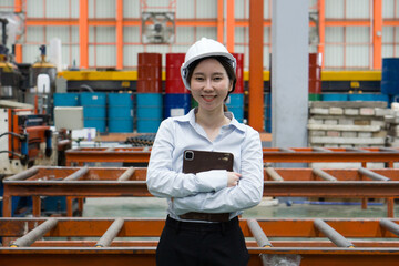 A dedicated female engineer dressed in safety gear stand smiling with arms crossed, holding tablet computer amidst a sprawling factory.