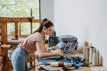 Young businesswoman working on laptop on workbench, standing by wooden loom. Upcycled textile,...