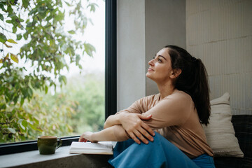 Hygge moment for beautiful woman sitting by a large window with a book. Woman is enjoying time for...