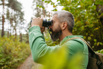 Handsome man watching wildlife with binoculars, enjoying peaceful atmosphere of forest, forestbathing