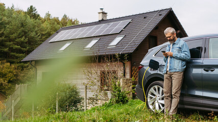 Man charging electric car in front of his house with solar panel system on roof behind him. Man...