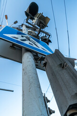 A low-angle perspective photograph of a pedestrian crossing signpost featuring traffic lights and electrical components against a blue sky. The image highlights urban infrastructure and city life