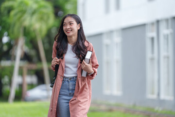 Portrait of a charming Asian female student in casual clothes. With backpack walking on campus her university Holding a book and smiling happily, enjoying the concept of people and education.