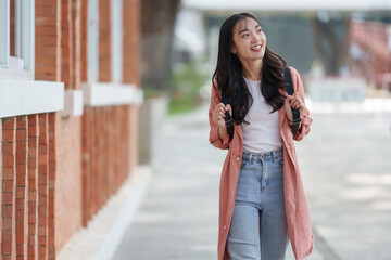 Educational concept Attractive, cool, Asian female student with backpack and notebook stands and smiles happily and relaxed. Outdoor learning atmosphere, university background, free space.