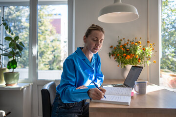 Concentrated middle aged woman works at home, planning day jotting down diary, comfortable sitting...