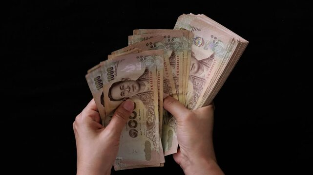 A woman's hands counting a large amount of Thai baht banknotes with a black background.