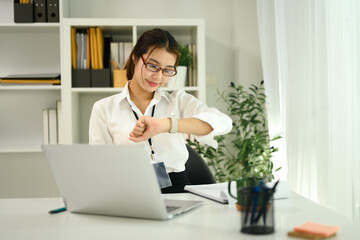 Asian female office worker sitting at her work desk and checking time on wristwatch